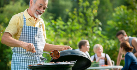 Older man cooking at BBQ with family in the background